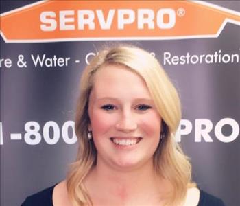 Female employee with blonde hair smiling in front of a SERVPRO black banner